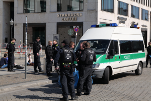 Eine Gruppe von Polizisten steht vor einem Starbucks-Café, rechts ein Van und links einige Menschen, im Hintergrund ein Gebäude mit Fenstern, einem Schild, einem Laternenmast und einem Zaun.