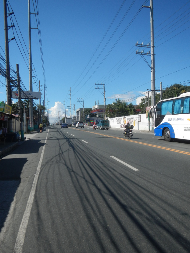 Ein Bus fährt auf einer Straße mit hohen Strommasten und Drähten, auf der linken Seite sind Geschäfte und Fußgänger zu sehen, sowie eine Wand, Bäume, Gebäude und ein bewölkter Himmel im Hintergrund.