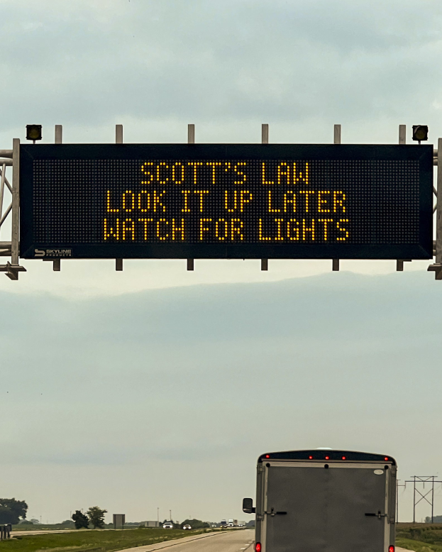 Ein großer Lkw fährt auf einer Autobahn mit einem Schild "Scott's Law: Schau es später nach, achte auf die Lichter" an der Seite, umgeben von Gras, Bäumen, Polen und einem sichtbaren Himmel.