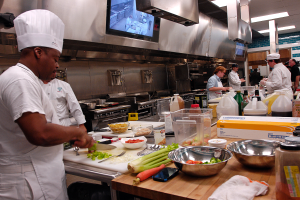 Eine Gruppe von Köchen in einer Küche, die Essen auf einem Schneidebrett zubereiten, mit einem Koch im Vordergrund in einer weißen Schürze und Mütze, der ein Messer hält und von Utensilien, Schüsseln, Gemüse, einem Handy, Flaschen und anderen Gegenständen umgeben ist, mit einem Fernseher und Deckenleuchten im Hintergrund.