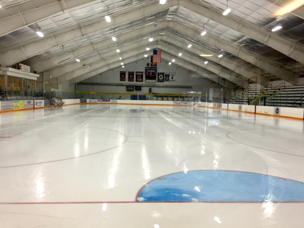 Große Indoor-Eisfläche mit Deckenbeleuchtung, Werbetafeln, einer Wand, Treppen mit Geländern und einigen Menschen, die einem Hockey-Sportplatz ähneln.