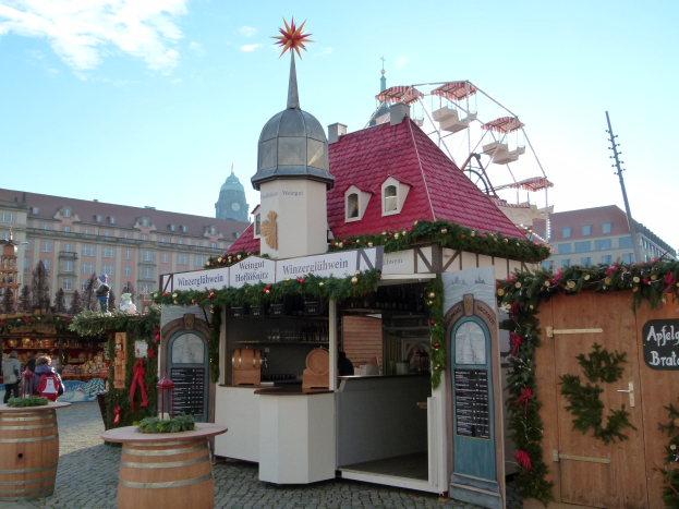 Ein kleines Gebäude mit einem roten Dach und einem Riesenrad davor, umgeben von Menschen und Festobjekten, mit Gebäuden, Bäumen und einem bewölkten Himmel im Hintergrund während des Oktoberfests in München, Deutschland.