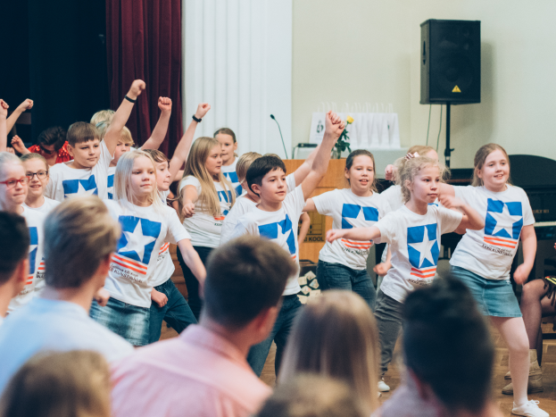 Eine Gruppe von Kindern in weißen Hemden und blauen Shorts tanzt auf einer Bühne vor einem sitzenden Publikum, mit einem Vorhang, Lautsprecher und einem Tisch mit Mikrofon im Hintergrund.