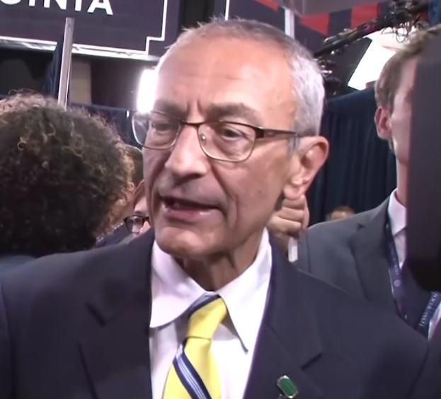 Ein Mann in Anzug und Krawatte spricht zu Reportern an einem Podium mit Mikrofon, umgeben von einer Menge, mit beleuchtetem Hintergrund und sichtbarem Schild.