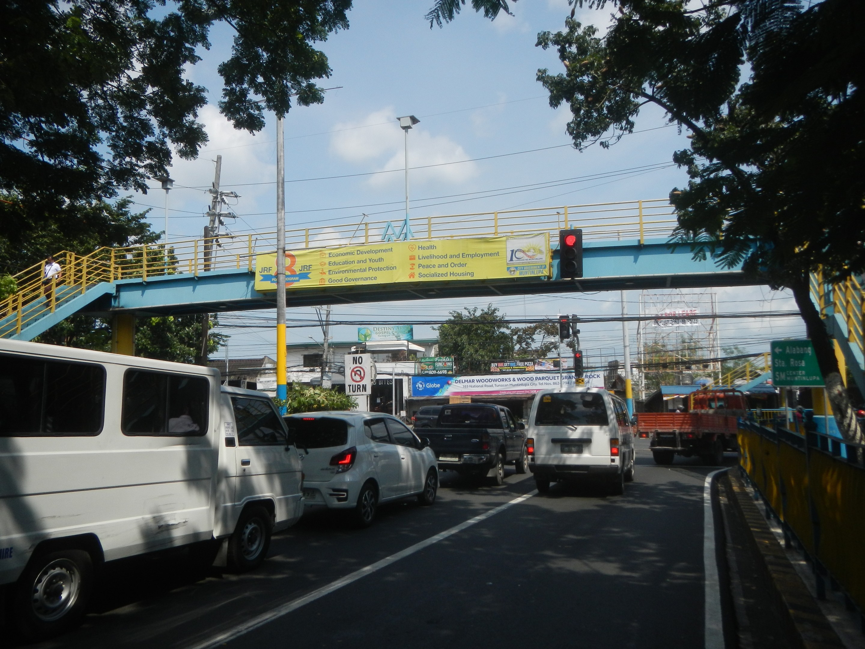 Bustling street scene with vehicles, a bridge with railings and steps, light poles, traffic signals, signboards, trees, buildings, and a cloudy sky.