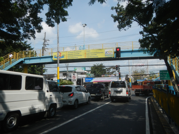 Bustling street scene with vehicles, a bridge with railings and steps, light poles, traffic signals, signboards, trees, buildings, and a cloudy sky.