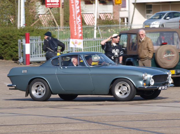 Eine Reihe von Autos mit Passagieren fährt auf einer Straße neben Gebäuden, Bäumen und verschiedenen städtischen Elementen, mit einer Straße unten.