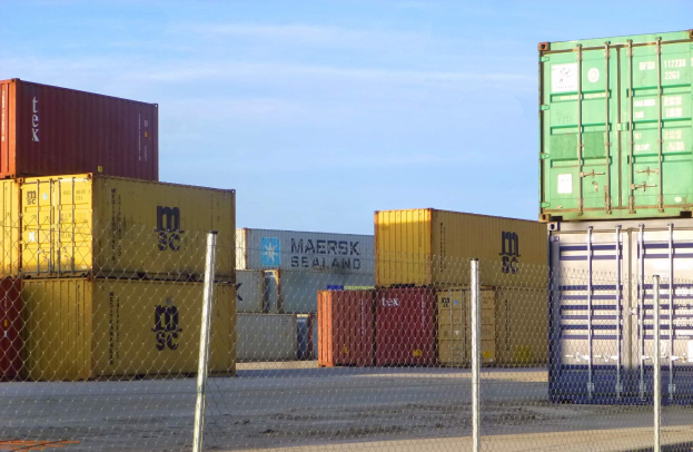 Ein hoher Stapel von Container hinter Maschendrahtzaun, mit einer Straße unten und einem bewölkten Himmel im Hintergrund.