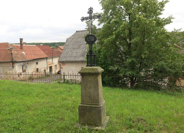Ein hölzernes Kreuz-Denkmal steht in einem von einem Metallzaun umgebenen Grasfeld, mit Bäumen, Häusern und einem bewölkten Himmel im Hintergrund, gewidmet den Holocaust-Opfern.