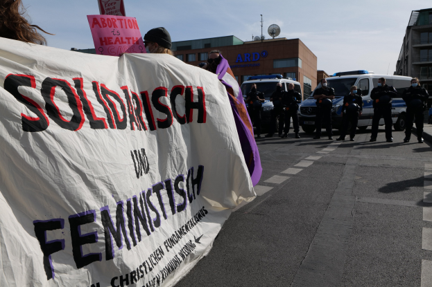 Gruppe von Menschen marschiert mit einem 'Solidarität und Feminismus'-Schild die Straße entlang, mit geparkten Fahrzeugen, Gebäuden, einer Schüsselantenne und einem klaren blauen Himmel im Hintergrund.