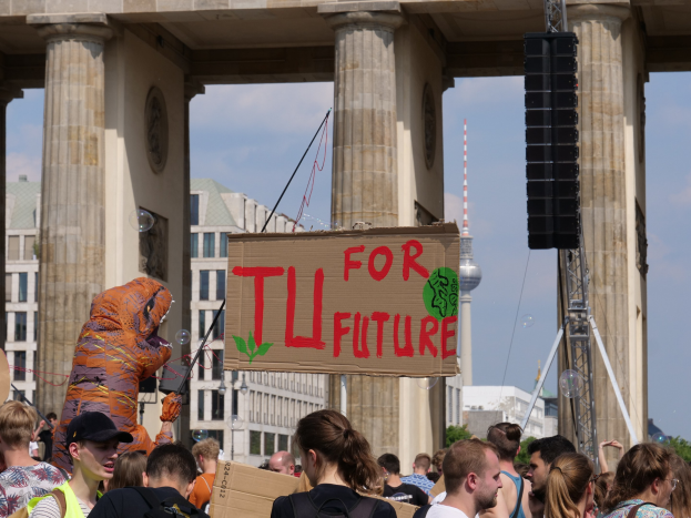 Eine Gruppe von Menschen steht vor dem Brandenburger Tor in Berlin, Deutschland, einige tragen Taschen und einer im Kostüm hält ein Schild, vor einem Hintergrund aus Gebäuden, Bäumen und einem Turm unter einem bewölkten Himmel.