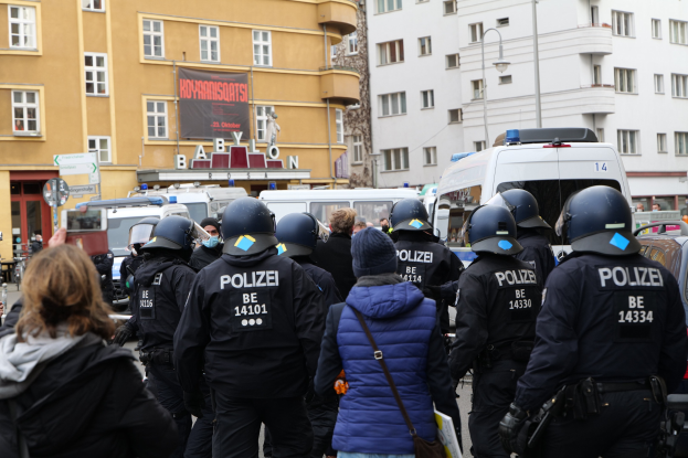 Polizeibeamte in Uniform vor einer Menge von Menschen mit Helmen und Jacken während einer Demonstration in Berlin, Deutschland, mit Fahrzeugen, Gebäuden, Laternen und einem Banner im Hintergrund.