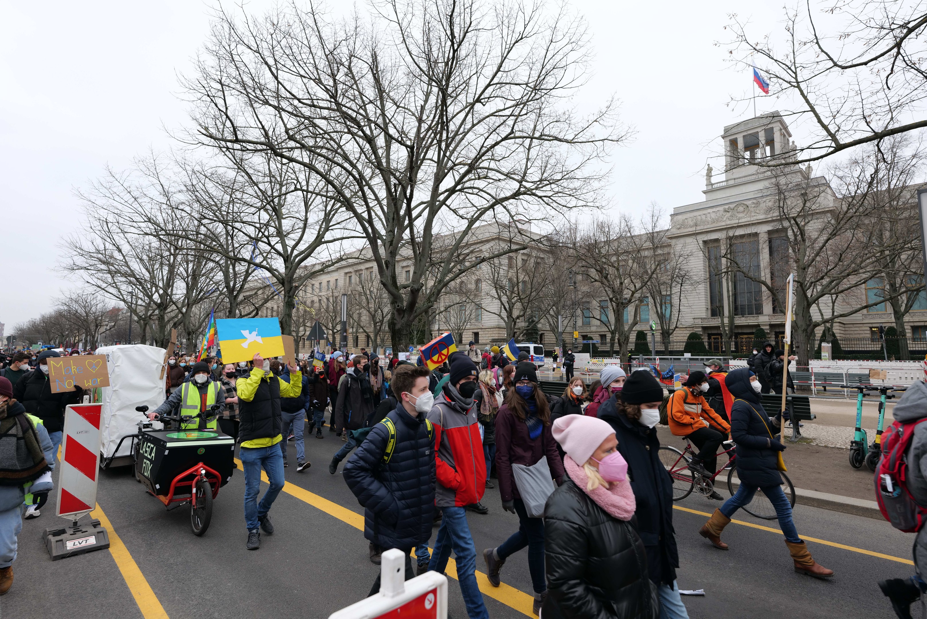 Eine große Gruppe von Menschen marschiert bei einer Demonstration auf einer Straße, einige halten Schilder und andere fahren Fahrräder, mit Bäumen und einem Gebäude im Hintergrund bei einem klaren blauen Himmel.