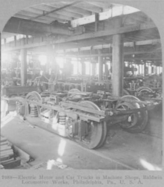 Schwarzes und weißes Foto eines Elektromotors und eines Lkw in einer industriellen Fabrikumgebung mit Maschinen, Säulen und einer sichtbaren Decke, einschließlich Text unten.