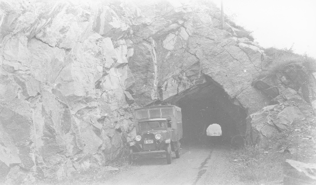 Schwarzes und weißes Foto eines Lastwagens, der durch einen Tunnel mit felsigen Wänden auf beiden Seiten fährt.