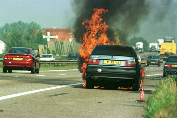 Ein Auto in Flammen auf dem Seitenstreifen, umgeben von anderen Fahrzeugen, mit B├Ąumen, Geb├Ąuden und einem klaren blauen Himmel im Hintergrund und einem Feuerl├Âscher auf der rechten Seite des Bildes.