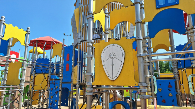 Ein Spielplatz mit buntem Spielgerät, einschließlich Stangen, Pfählen und einem Schirm, einige Menschen, Bäume und einen klaren blauen Himmel im Hintergrund.