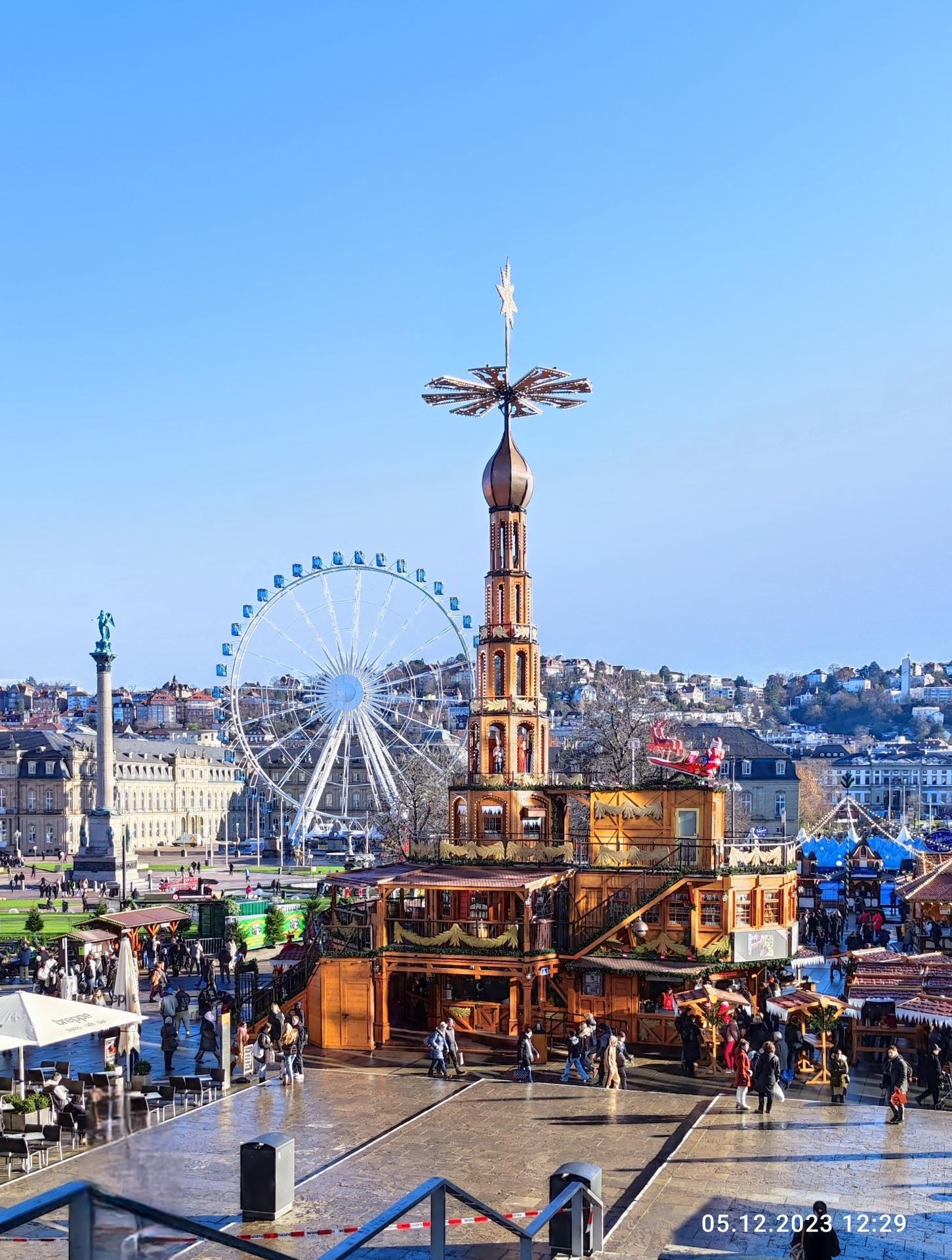 Ein belebter Stadtplatz mit einem großen Riesenrad in der Mitte, umgeben von Gebäuden, Bäumen und einem klaren blauen Himmel, mit Menschen auf Stühlen sitzend, stehend und gehend, sowie Zelten, Geländern und anderen Gegenständen und einem Wasserzeichen unten.