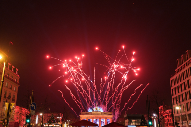 Eine belebte Stadtstraße an einem Neujahrsabend in Berlin, mit Gebäuden, Bäumen, Laternenmasten, Verkehrsampeln, Schildern, Zelten, Menschen und einem prächtigen Feuerwerk am Himmel.
