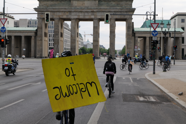 Eine Gruppe von Radfahrern fährt am Brandenburger Tor in Berlin vorbei, einer hält ein gelbes Schild, mit Laternenpfählen, Verkehrsampeln, Gebäuden, Bäumen und einem klaren Himmel im Hintergrund.