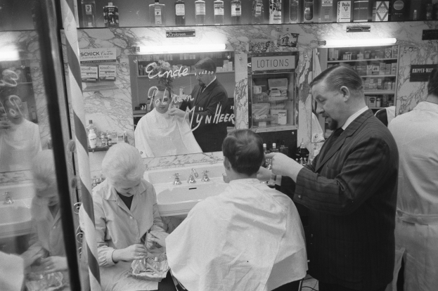 Schwarz-weißes Foto eines Mannes, der einer Frau im Friseursalon die Haare schneidet, mit einem Waschbecken, Spiegel und Regalen mit Vorräten im Hintergrund.