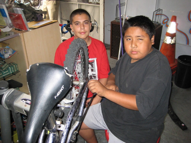 Zwei Personen reparieren ein Fahrrad in der Mitte, mit einem Schreibtisch und verschiedenen Gegenständen links und Fahrrädern, Ersatzteilen und anderen Gegenständen im Hintergrund.