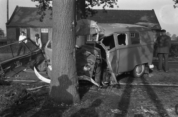 Schwarzes und weißes Bild eines auf der Straße gestrandeten Autos mit einem Baum im Vordergrund, einigen Menschen in der Nähe und Häusern, Bäumen und einem Mast im Hintergrund.