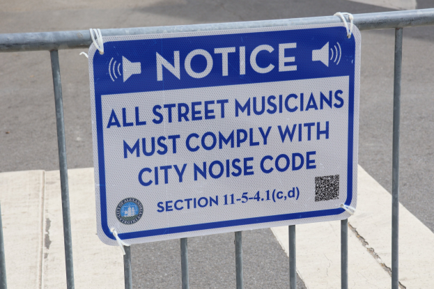 Anschlagtafel an einem Zaun mit einem Schild, das Straßenmusikern den Einhaltung der städtischen Lärmvorschriften vorschreibt, Straße im Hintergrund sichtbar.