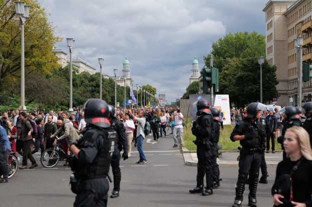 Eine Gruppe von Menschen geht eine Straße entlang mit Polizisten in Helmen, die Waffen halten, Fahrrädern, Laternen, Verkehrsampeln, Bäumen, Gebäuden mit Fenstern und einem bewölkten Himmel im Hintergrund.