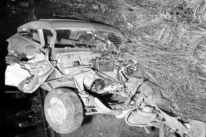 Ein Schwarz-Weiß-Foto eines schwerbeschädigten Autos am Straßenrand, mit einer eingedrückten Front, zersplitterten Fenstern, einem Baum im Hintergrund und verstreuten Gegenständen an der Unfallstelle.
