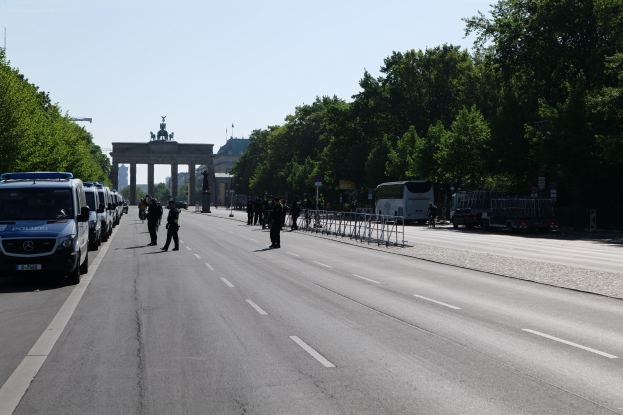 Polizeifahrzeuge entlang einer Straße mit Barrieren vor dem Brandenburger Tor geparkt, Menschen gehen, Bäume und Statuen im Hintergrund unter einem sichtbaren Himmel.
