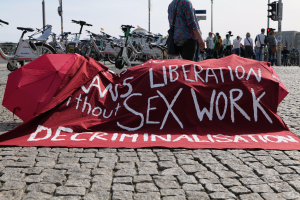 Gruppe von Menschen mit Schirmen in der Nähe von Fahrrädern mit einem roten Protesttuch, auf dem "Befreiung ohne Entkriminalisierung der Sexarbeit" steht, auf dem Bürgersteig stehend, umgeben von städtischen Elementen wie Ampeln und Gebäuden.