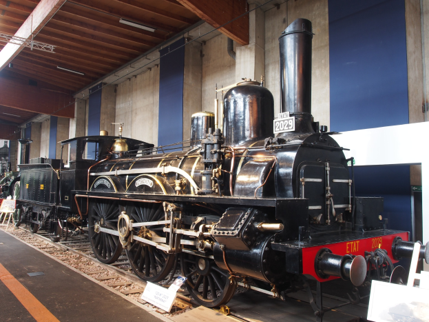 Alte Dampflokomotive in einem Museum ausgestellt, umgeben von Eisenbahnschienen und informativen Schildern, mit einer Wand und Deckenbeleuchtung im Hintergrund.