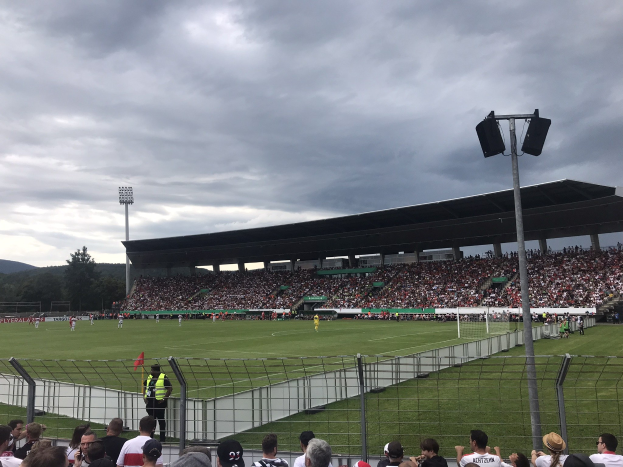 Große Zuschauermenge bei einem Fußballspiel in einem bewölkten, offenen Stadion mit beleuchtetem Feld, eingezäuntem Rand, Laternenmasten und fernen Hügeln.