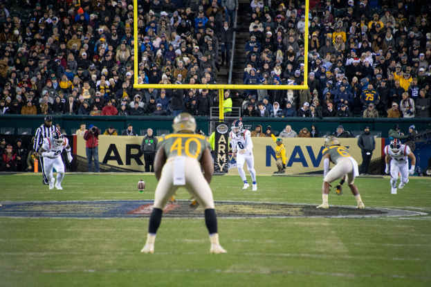Fußballspieler in Sportkleidung und Helmen konkurrieren auf einem Feld mit Zuschauern im Hintergrund, Stadionstrukturen und Textanzeigetafeln sind sichtbar.