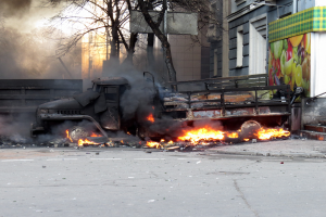 Ein Lkw brennt am Straßenrand, mit Rauch, der aus ihm aufsteigt, während Bäume, Gebäude und ein klarer Himmel im Hintergrund zu sehen sind.