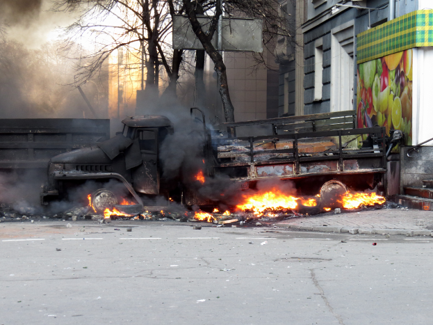 Ein Lkw brennt am Straßenrand, mit Rauch, der aus ihm aufsteigt, während Bäume, Gebäude und ein klarer Himmel im Hintergrund zu sehen sind.