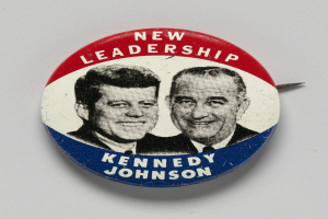 Ein Ansteckbutton auf einer weißen Oberfläche zeigt zwei Bilder von Kennedy und Johnson mit dem Text "Neue Führung" darunter.