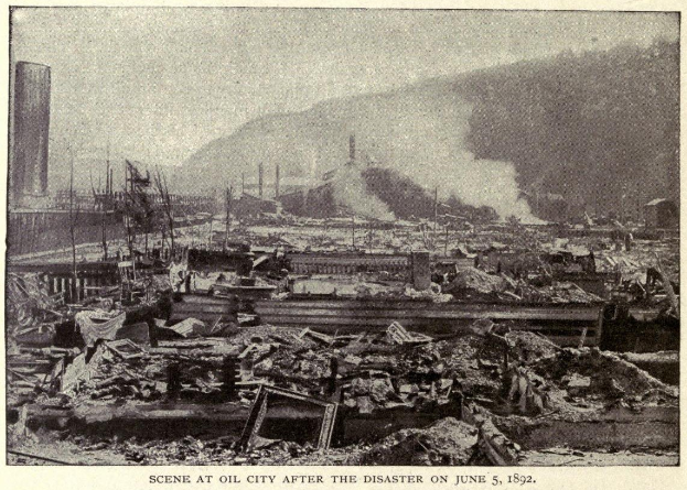 Schwarzes und weißes Foto einer Stadt nach einer Katastrophe am 5. Juni 1892, das Gebäude, Pfähle, Rauch, einen Hügel im Hintergrund und Text am Boden zeigt.