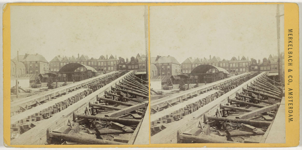Schwarz-weiß-Foto einer Bahntrasse mit Gebäuden im Hintergrund, Text 'Merkelbach & Co, Co. Amsterdam' rechts und verstreuten Holzobjekten im Vordergrund.