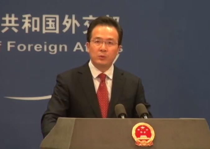 Mann im Anzug und Krawatte spricht an einem Podium mit Mikrofonen, Podiumslogo und Banner im Hintergrund.