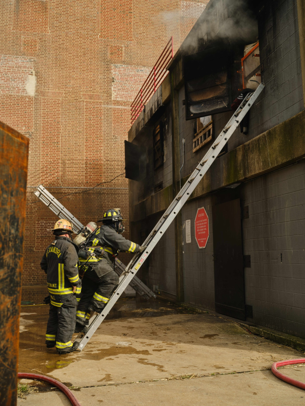 Feuerwehrleute mit Helmen klettern an einer Leiter zu einem raucherfüllten Gebäude hoch, während in der Nähe ein metallisches Objekt und Rohre zu sehen sind.