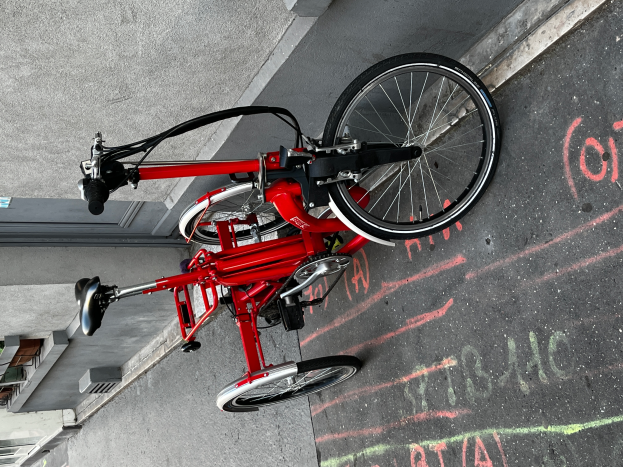 Ein rotes Klappfahrrad, das auf der Seite einer Straße geparkt ist, mit einem Gebäude im Hintergrund, das Text trägt.