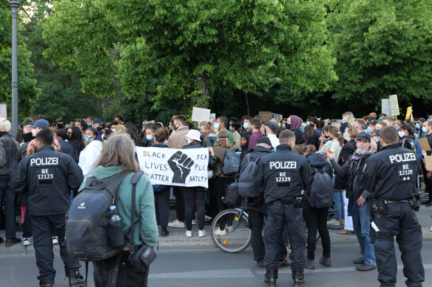 Eine große Gruppe von Menschen nimmt an einer Black Lives Matter Demonstration in Berlin teil, einige halten Schilder und andere tragen Kappen und Taschen, vorne steht ein Fahrrad und im Hintergrund sind Bäume und ein Pfahl zu sehen.
