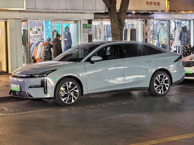 Ein Mitsubishi Lancer EV Elektroauto auf einer nächtlichen Stadtstraße geparkt, umgeben von Gebäuden, beleuchteten Schaufenstern, Schaufensterpuppen in Kleidern und verschiedenen Schildern.