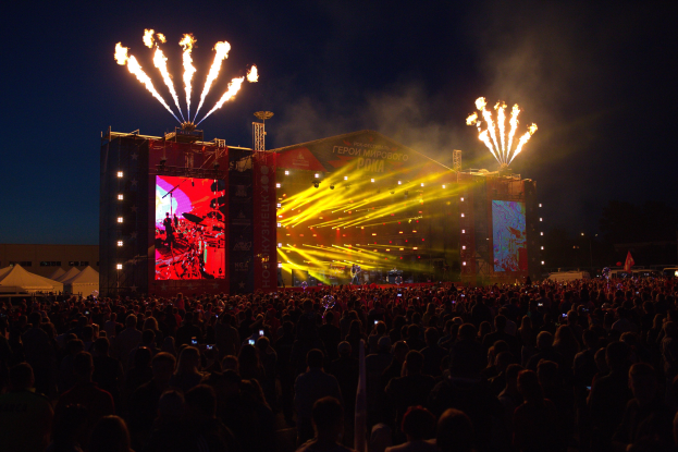 Eine große Menschenmenge schaut bei einem Musikfestival auf eine hell erleuchtete Bühne, mit Zelten links und Feuerwerk am dunklen Himmel dahinter.