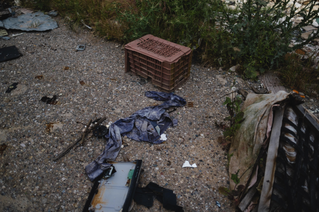 Haufen Müll einschließlich einer Plastikbox und Kleidung auf der Seite einer Straße in der Nähe eines Zaunes, mit Pflanzen und Gras im Hintergrund.