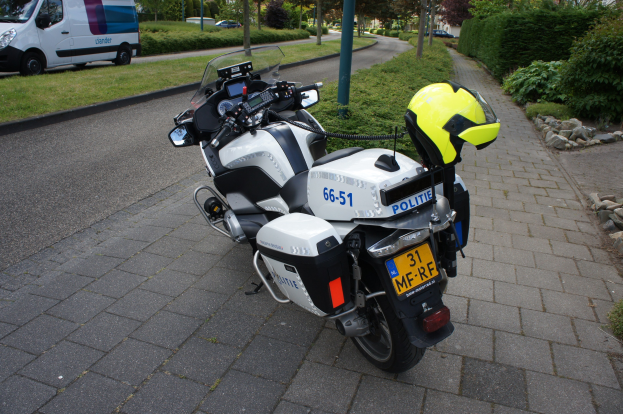 Ein auf der Straße geparktes Polizeimotorrad mit einem Helm darauf, umgeben von Bäumen, Polen, Gebäuden, Pflanzen, Gras, Steinen und anderen Fahrzeugen.