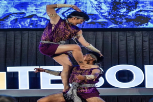 Zwei Akrobaten führen auf der Bühne eine Darbietung auf, der Mann hält die Frau in seinen Armen, während beide purpurfarbene Kleider tragen, mit Publikum und Bildschirm im Hintergrund.