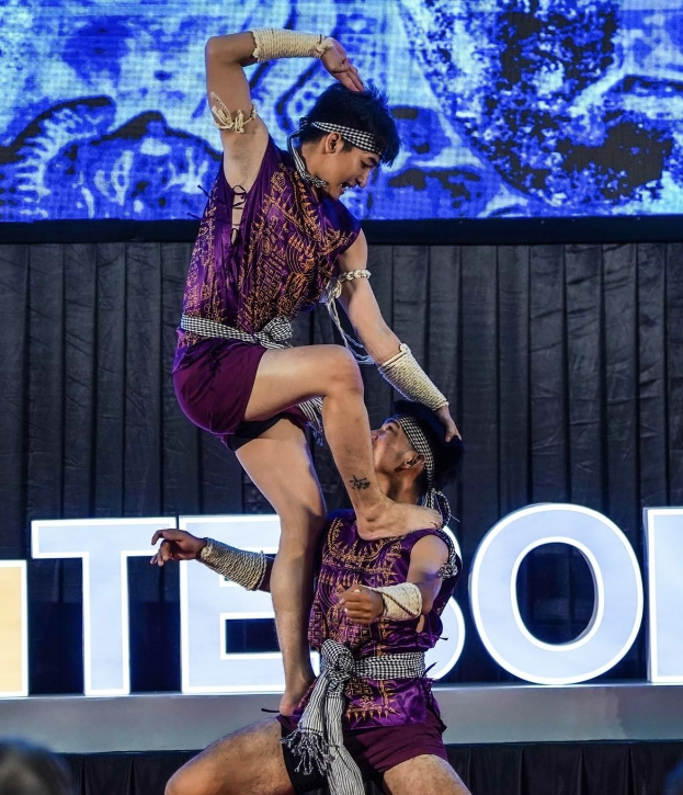 Zwei Akrobaten führen auf der Bühne eine Darbietung auf, der Mann hält die Frau in seinen Armen, während beide purpurfarbene Kleider tragen, mit Publikum und Bildschirm im Hintergrund.